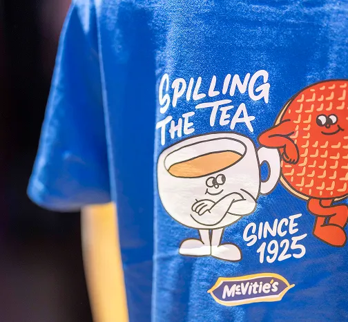Blue McVitie's T-shirt featuring a cartoon chocolate digestive and cup of tea with the slogan “Spilling the tea since 1925.”
