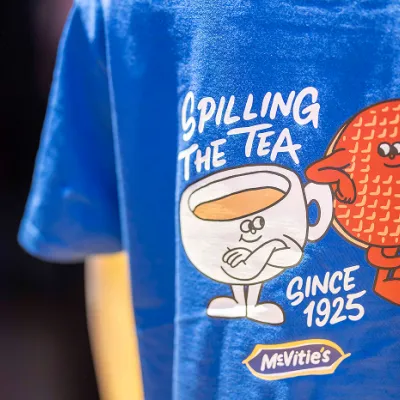 Blue McVitie's T-shirt featuring a cartoon chocolate digestive and cup of tea with the slogan “Spilling the tea since 1925.”