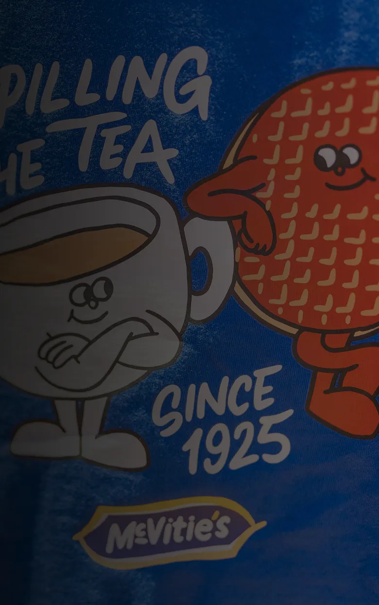 Blue McVitie's T-shirt featuring a cartoon chocolate digestive and cup of tea with the slogan “Spilling the tea since 1925.”